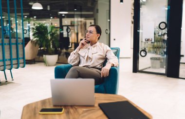 Genç bayan çalışan, gözlüklü ve şık giysili. Dizüstü bilgisayarlı, bacak bacak üstüne atmış bir şekilde modern iş merkezinde düşünceli bir şekilde oturuyor.