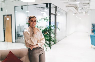 Modern ofiste rahat bir kanepeye yaslanırken başparmağıyla çeneye dokunup gülümseyen gündelik kıyafetli genç bir kadın.