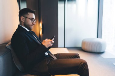 Resmi takım elbiseli, gözlüklü ciddi bir erkek girişimcinin koltukta oturması ve ofisten çıkarken cep telefonunu karıştırması.