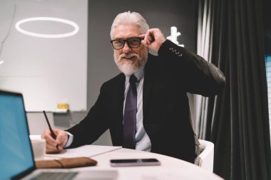 Resmi olarak yaşlı girişimci, gözlük ayarlarken gülümseyip kameraya bakarken ve bilgisayarlı akıllı telefon defteriyle masada otururken ofisteki iş yerindeki not defterine notlar alırken giyer.