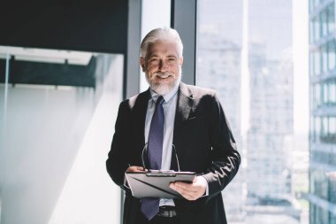 Zarif takım elbiseli, gülümseyen ve elinde pano gözlüklerle cam duvarın yanında duran yaşlı bir işadamı gündüz vakti bulanık şehir binalarına karşı kameraya bakarken görülüyor.