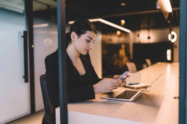 Modern ofiste dizüstü bilgisayar ve kalemle çalışırken resmi giyinmiş, cep telefonu ekranı seyreden ve gezinen genç ve sakin iş kadınının yan görüntüsü