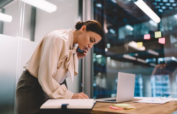 Вид сбоку на сконцентрированную молодую предпринимательницу, разговаривающую по мобильному телефону, за столом с ноутбуком рядом с окном в офисе