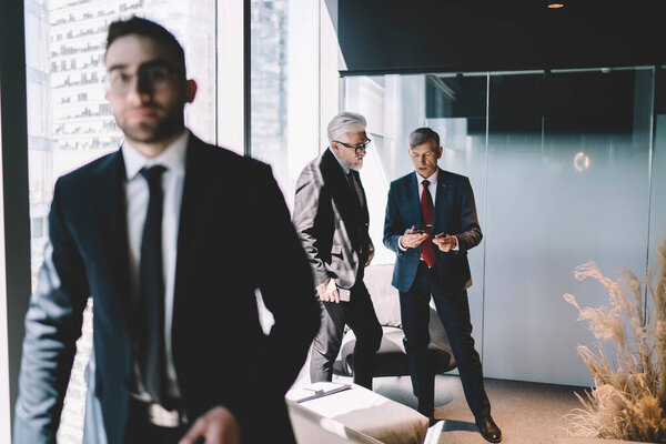 Размытый человек, входящий в офис с различными старшими бизнес-партнерами на заднем плане, используя мобильный телефон и разговаривая в современном офисном здании