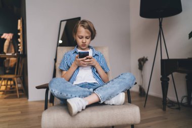 Sıradan giysiler içinde vücut çocuğu koltuğunda bağdaş kurup hafta sonları akıllı telefondan evdeki oturma odasında video izliyor.