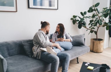 Genç çift kanepede dinleniyor ve birbirlerine akıllı telefonlarıyla bakıyorlar ve birlikte oturma odasında cep telefonlu kahve fincanlarıyla vakit geçiriyorlar.