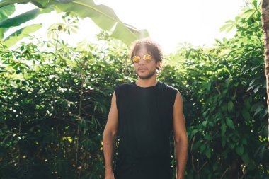 Siyah gömlekli, güneş gözlüklü, yeşil ağaçların yanında duran ve gündüz vakti Endonezya 'nın parlak güneşinde kameraya bakan kendinden emin genç bir erkek.