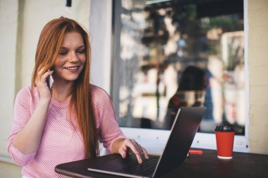 Dizüstü bilgisayarda çalışan ve kafeteryadaki masada otururken akıllı telefondan konuşan zarif pembe kazaklı, serbest çalışan genç bir kadın.