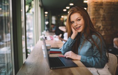 Boş zamanlarında kafeteryada bilgisayarlı akıllı telefon günlüğü ile ahşap masada oturup gülümserken ve masaya yaslanırken kameraya bakıp, yanağına dokunan ve masaya yaslanan genç, pozitif bir serbest kadın.