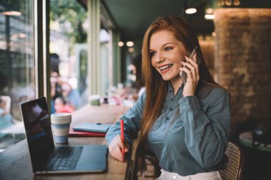 Modern kafeteryada bir fincan kahveyle otururken akıllı gündelik kıyafetlerle konuşan ve bilgisayarla konuşan neşeli, genç bir bayan serbest yazar.