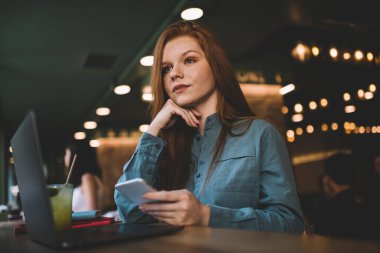 Düşük açılı kadın serbest çalışan, parmakları çeneye değen, akıllı telefonu elinde tutan ve kafedeki molada dizüstü bilgisayarlı cam günlükle masada oturan.
