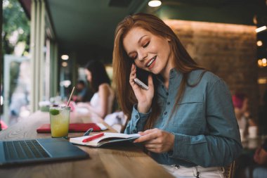 İyimser gülümseyen, şık giyinmiş, akıllı telefonlu bir kadın. Kafede dizüstü bilgisayar ve gündüz vakti bir bardak meyve suyu içerken.
