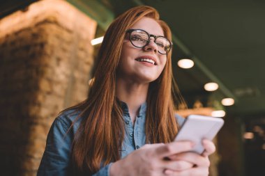 Düşük açılı, yansıtıcı gözlüklü, gülümseyen ve başka yöne bakan genç bir kadın. Gün ışığında aydınlatmalı, bulanık bir kafede zaman geçirirken akıllı telefon kullanıyor.