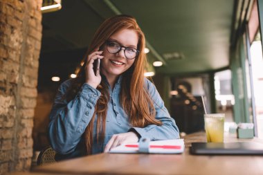 Düşük açılı, gözlüklü, şık giyinen, akıllı telefondan konuşan, elinde not defteri ve bir bardak meyve suyu olan bir kadın.