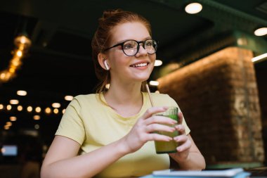 Kafeteryada oturup müzik dinlerken, elinde bir bardak taze içecek tutan, gündelik giysiler ve gözlüklerle, iyimser, genç bir bayan serbest yazar.