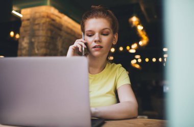 Genç serbest çalışan bir bayan bilgisayarla masada otururken ve gece bulanık ve aydınlanmış bir kafeden ekrana bakarken telefonla konuşuyor.