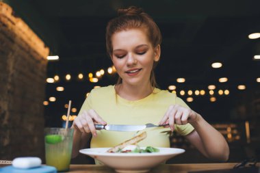 Genç ve mutlu bir bayan elinde bir kase meyve suyu ve bulanık kafede yemek yerken gülümseyerek masaya bakıyor.