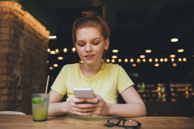 Olumlu genç bir bayan gülümsüyor ve ekrandaki cep telefonuna bakıyor. Bu arada ahşap masada otururken ve bir bardak meyve suyu gözlüğüyle bulanık bir kafede.