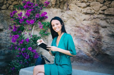Gülümseyen genç İspanyol kadın, taş bir bankta oturmuş çiçeklerin yanında bacak bacak bacak üstüne atmış ve parkta roman okurken kameraya bakıyor.