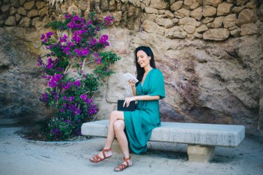 Yandan gülümseyen İspanyol bir kadının taş bankta bacaklarının çapraz durduğu çiçeklerin yanında oturup kitap okurken ekrandaki cep telefonu ekranına bakması.