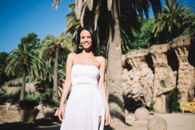 Beyaz elbiseli neşeli genç bayan kumlu sahilde dururken gülümseyip kameraya bakıyor ve tatil boyunca yaz gününün tadını çıkarıyor.