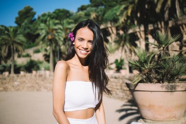 Beyaz elbiseli neşeli genç bir bayan saçları çiçekli kameraya bakıyor ve yaz tatilinde kumlu sahilde dinlenirken gülümsüyor.