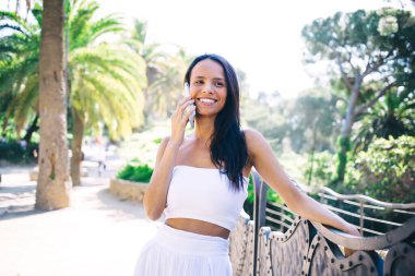 Beyaz günlük giysiler giyen, cep telefonuyla konuşurken gülümseyen ve yaz tatilinde tropik tatil beldelerinde dinlenen etnik bir kadın.