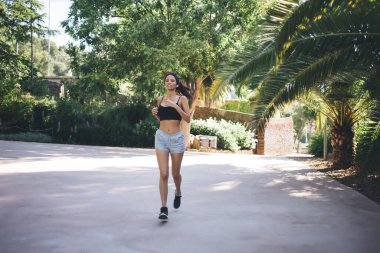 Spor ayakkabılı, yeşil tropikal ağaçların yanında koşan ve yaz günlerinde tatil köyünde koşu egzersizi yaparken kameraya bakan bir sürü İspanyol kadın.