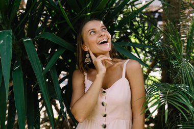 Hafif yaz elbisesi içinde neşeli genç bir bayan. Tatilde tropikal egzotik bahçede dikilirken gülümsüyor ve başka tarafa bakıyor.