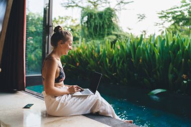 Sütyen ve beyaz pantolonlu, villa terasında oturan ve tatilde uzaktan çalışırken dizüstü bilgisayarında bilgi yazan pozitif kadın serbest yazar.