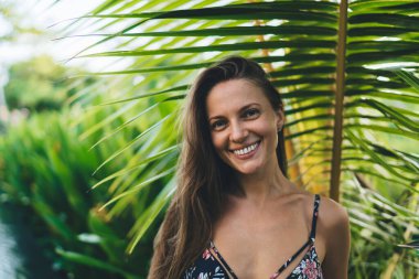 Mayo giymiş hoş kahverengi saçlı bayan gün ışığında tatil sırasında yeşil tropikal bitkilerin bulanık arka planına karşı kameraya bakıyor.