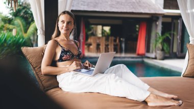 Şık giyinmiş, şilte ve dizüstü bilgisayarda oturmuş tatil köyünde uzak bir projede çalışırken yüzme havuzunun yakınına bakan pozitif, genç bir kadın serbest yazar.