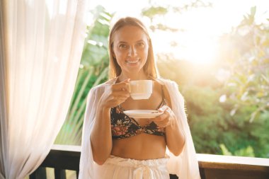 Sıcak kahve fincanı ve çay tabağıyla pencerenin yanında güneş ışığında bulanık yeşil parka bakarken şık giyinmiş gülümseyen genç bir kadın kameraya bakıyor.