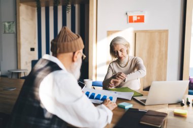 Odaklanmış yaşlı kadın dizüstü bilgisayarlı not panosuyla masada oturuyor ve üst düzey erkek ofiste beklerken ve diyagramları kontrol ederken saati kontrol ediyor.
