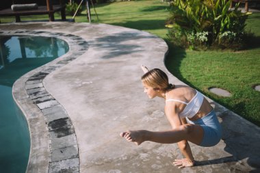 Havuz kenarında zor yoga yapan, elleri yerde, bacakları havada esneyen ve güneşli yaz parkında yan yana duran genç ve formda bir kadının yan görüntüsü.