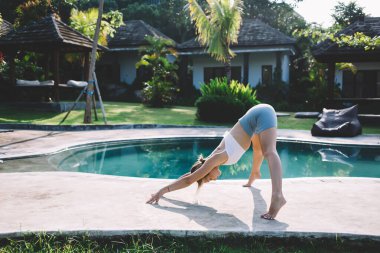 Güneşli bir günde yeşil parkta, parlak güneş ışığı altında Prasarita Padottasana yogası yaparken geniş bacaklı ve öne doğru eğilmiş bir kadının yan görüntüsü.