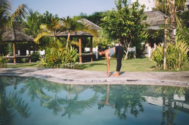 Yüzme havuzu yakınlarındaki güneşli tropikal tatil beldesinde yoga yaparken spor yapan aktif giyimli formalı bir kadının yan görüntüsü.