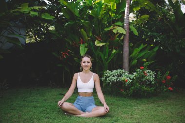 Spor kıyafetleri içinde genç bir kadın. Parktaki yeşil çimlerde meditasyon yaparken gülümsüyor ve kameraya bakıyor.