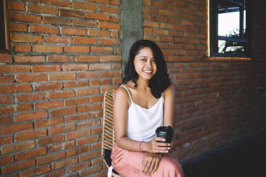 İyimser etnik kadın kameraya bakıp gülümserken bir yandan da elinde sıcak içecekle tuğla duvara yaslanıyor.