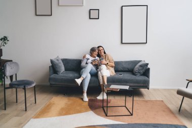 Mutlu genç kız huzur dolu bir atmosferde bacak bacak bacak üstüne atmış gri kanepede dinlenirken yaşlı anneye sarılıyor.