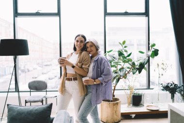 Gülümseyen kız ve anne gündelik kıyafetleriyle kucaklaşırken modern apartmanın penceresinde kameraya bakarak sıcak içecekler içiyorlar.