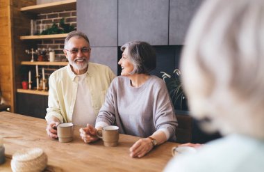 Gözlüklü sakallı adama bakan yaşlı bir kadın sıcak içecekler içerken masada oturuyor ve tanınmayan arkadaşlarıyla harika vakit geçiriyor.