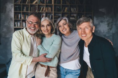 Bir grup pozitif orta yaşlı insan gülümseyip kameraya bakarken birbirlerine sarılıyorlar ve evdeki oturma odasında başkalarına el kaldırıyorlar.