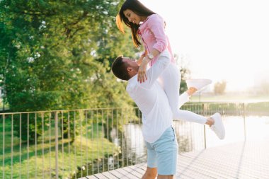 Sevgili sevgilinin, yaz parkında göletin kenarında dikilip birbirine bakarken etnik kız arkadaşını yerden kaldırması.