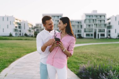 Mutlu genç erkek arkadaş kız arkadaşına polen çiçeği hediye ediyor cep telefonu kullanıyor ve şehir parkında asfalt yolda dururken gülümsüyor.