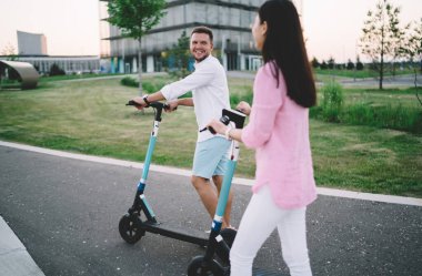 Neşeli adam ve kadının günlük kıyafetleri içinde asfalt yolda elektrikli scooterlarla yürümeleri ve hafta sonu boyunca birbirlerine bakmaları.
