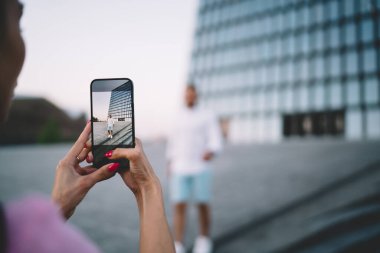 Kırmızı manikürlü, elinde cep telefonu olan tanınmamış bir kadın yazın günbatımında şehir caddesinde modern cam bina yakınlarında fotoğraf çekiyor.