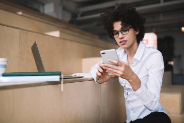 Milenyum Kafkas kızı iş sırasında akıllı telefon kullanıyor ve izliyor. Modern başarılı kadın kavramı. Gözlüklü genç bir kadın çalışma odasındaki ahşap bankta oturuyor.