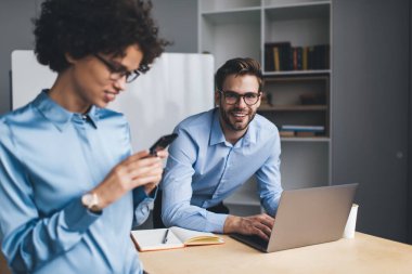 Masada dizüstü bilgisayarda çalışan gülümseyen iş adamı ve bayan meslektaşı cep telefonu kullanırken kameraya bakıyor. Modern başarılı insanlar kavramı. Genç Kafkasyalı milenyum meslektaşları görev başında