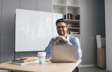 Milenyum Kafkas işadamı masadaki dizüstü bilgisayarda çalışırken kameraya bakıyor. Modern başarılı adam kavramı. Geniş ofiste gözlüklü ve resmi takım elbiseli genç adam.